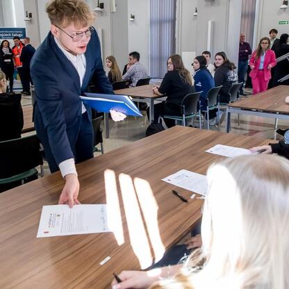 Wojewódzki konkurs wiedzy o pierwszej pomocy, uczestnicy siedzą przy stołach, rozdzielany lub zbierany papier. (wygenerowano przez AI)