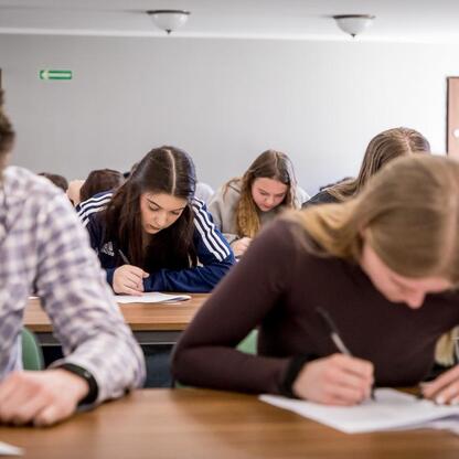 Klasa uczestników konkursu wiedzy o pierwszej pomocy, siedzący przy drewnianych biurkach, skupieni na testach. (wygenerowano przez AI)