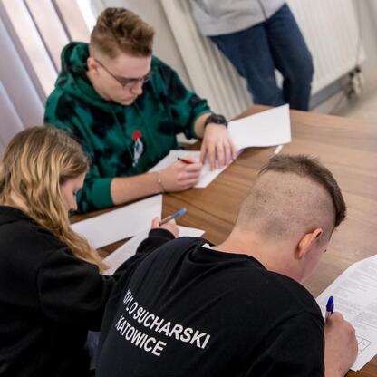 Grupa studentów wzięła udział w finałowym etapie konkursu wiedzy o pierwszej pomocy organizowanego przez Śląskie Youth Parliament. Uczestnicy, ubrani w casual, skupieni są na pisaniu testu. (wygenerowano przez AI)