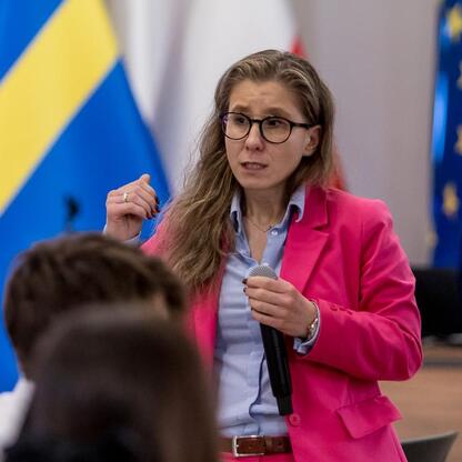 Kobieta w różowej kurtce przemawia do mikrofonu, podczas finału konkursu pierwszej pomocy województwa śląskiego. (wygenerowano przez AI)