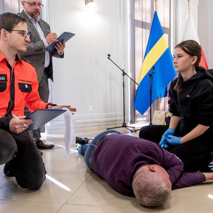 Paramedic P. Nowicki demonstruje techniki reanimacji na manekinie, wspomagany przez uczestniczkę konkursu. (wygenerowano przez AI)