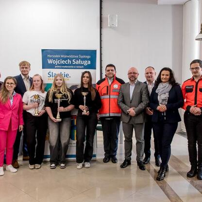 Zakończenie konkursu wiedzy o pierwszej pomocy województwa Śląskiego, uczestnicy w białych mundurach i czerwonych kamizelkach otrzymują trofea. (wygenerowano przez AI)