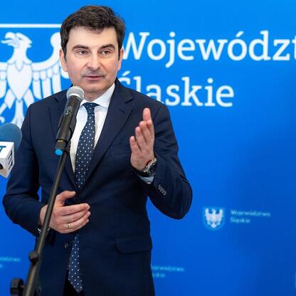 Mężczyzna w ciemnym garniturze przemawia na podium podczas konferencji prasowej Województwa Śląskiego. (wygenerowano przez AI)
