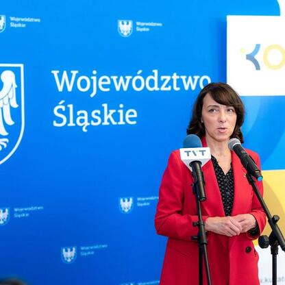 Kobieta w czerwonej kurtce przemawia na podium podczas konferencji prasowej dotyczącej Kongresu Oświaty Śląskiego. (wygenerowano przez AI)