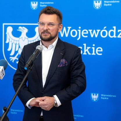 Mężczyzna w ciemnym garniturze i białej koszuli przemawia na podium z mikrofonami, podczas konferencji prasowej w Województwie Śląskim. (wygenerowano przez AI)