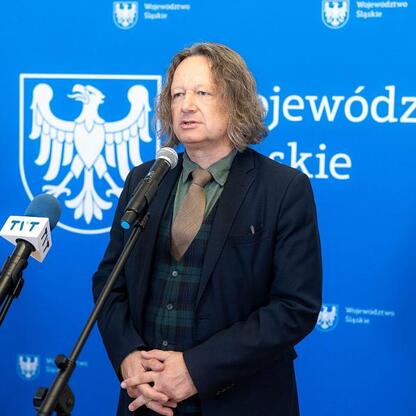 Mężczyzna w ciemnym garniturze, zielonym koszulą i krawatem w kratę, przemawia na podium w Urzędzie Marszałkowskim Województwa Śląskiego, podczas konferencji prasowej związnej z podpisaniem umowy doty (wygenerowano przez AI)