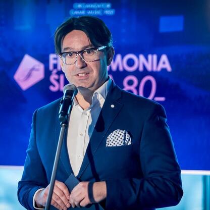 Mężczyzna w ciemnym garniturze z mikrofonem, prezentuje Game Music Fest w Filharmonii Śląskiej. (wygenerowano przez AI)