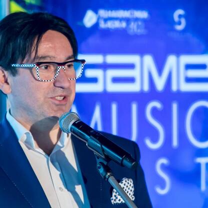 Mężczyzna w garniturze przemawia w mikrofonie podczas konferencji prasowej Game Music Fest, z logo Filharmonii Śląskiej na tle. (wygenerowano przez AI)