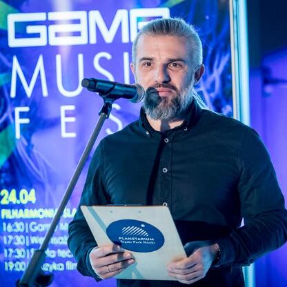 Na podium z mikrofonem stoi organizator festiwalu Game Music Fest, który rozpoczyna konferencję prasową. (wygenerowano przez AI)