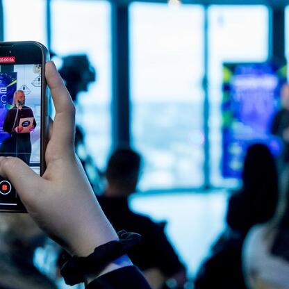 Fotografia z festiwalu Game Music Fest, gdzie uczestnik nagrywa prezentację na smartfonie, z tłem kolorowego, zrzeszonego tłumu. (wygenerowano przez AI)