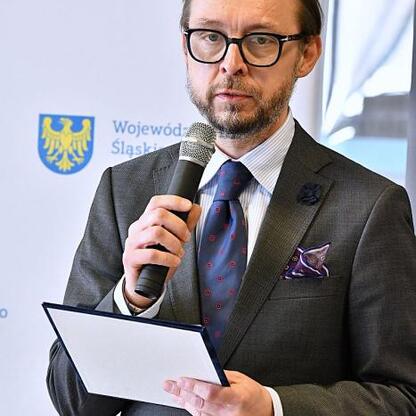 Na zdjęciu: Mężczyzna w garniturze przemawia na konferencji prasowej w Katowicach, prezentując projekt Modern Lib. (wygenerowano przez AI)