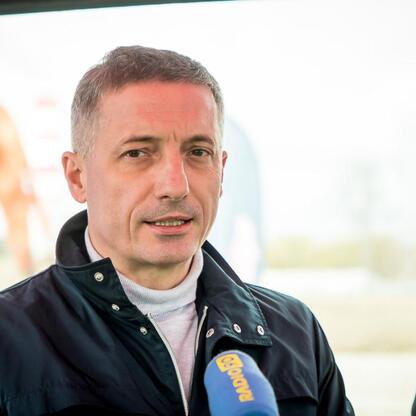 Widoczny na zdjęciu mężczyzna w czarnej kurtce i białej koszulce, mówiący w mikrofonie z napisem 