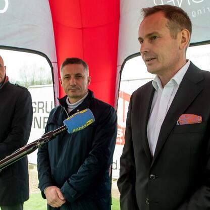 Trzech mężczyzn w garniturach stoi przed czerwono-białym namiotem, uczestnicząc w uroczystości symbolicznego wmurowania kamienia węgielnego nowego centrum materiałowego w Rybniku. (wygenerowano przez AI)