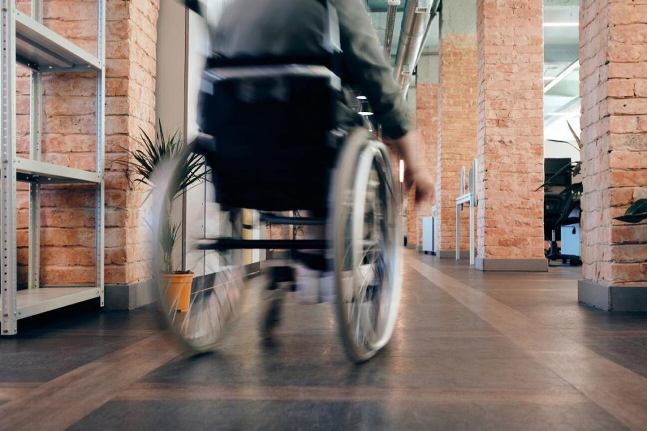 Osoba w wózku inwalidzkim porusza się po nowoczesnym biurze z ceglanymi ścianami i szafami w stylu przemysłowym, podkreślając temat dostępności i inkluzji. (wygenerowano przez AI)