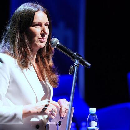 Kobieta w białym płaszczu przemawia na konferencji o transformacji energetycznej, gestykuluje, mikrofon w ręce. (wygenerowano przez AI)