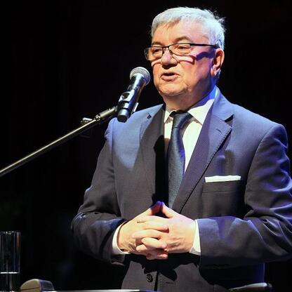 Mężczyzna w ciemnym garniturze i krawacie stoi na podium z mikrofonem, przemawia na konferencji o transformacji energetycznej miast. (wygenerowano przez AI)