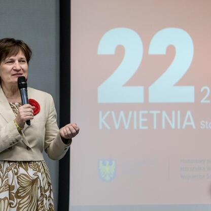 Podczas II Posiedzenia Rady Młodzieżowej na Stadionie Śląskim, kobieta w beżowym garniturze przemawia do publiczności, podkreślając znaczenie aktywności młodzieży. (wygenerowano przez AI)