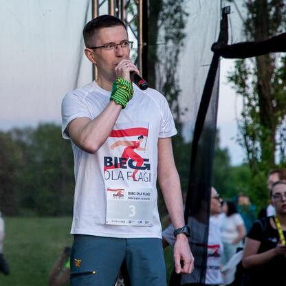 Na Biegu dla Flagi 2025 w Parku Śląskim mąż w białej koszulce z czerwono-białym wzorem przemawia do publiczności. (wygenerowano przez AI)