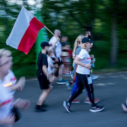 Zawodnicy biegną w Parku Śląskim, jedna z nich macha flagą Polski, symbolizując patriotyzm. (wygenerowano przez AI)