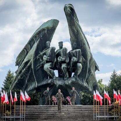 Słup pomnikowy, flagi, apel poległych, kwiaty, hołd zwycięzcom. (wygenerowano przez AI)