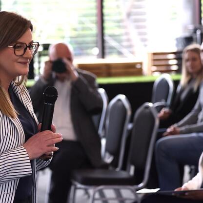 W czasie forum biznesowego w Arenie Zabrze, kobieta w pasiastym płaszczu przemawia do publiczności w konferencyjnym pomieszczeniu. (wygenerowano przez AI)