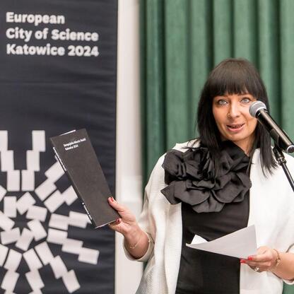 Podczas Forum Przyszłości Miasta Nauki w Katowicach na Uniwersytecie Śląskim kobieta przemawia na podium, trzymając książkę i papier. (wygenerowano przez AI)