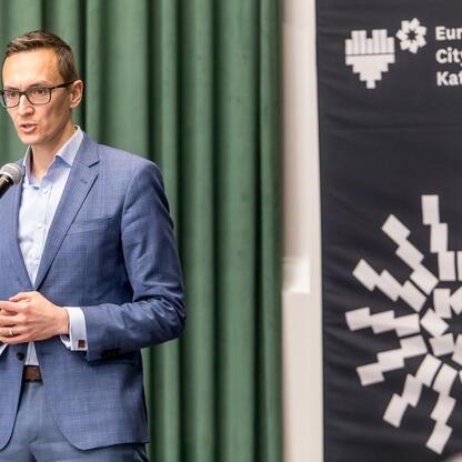 Mężczyzna w niebieskim garniturze i okularach przemawia na podium podczas konferencji 