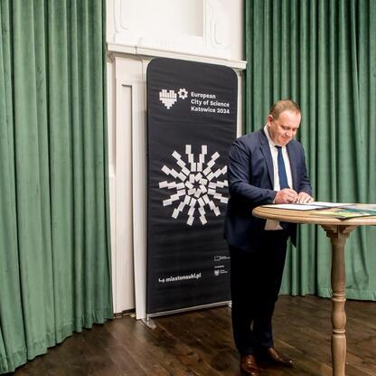 Na Uniwersytecie Śląskim podczas obchodów Europejskiego Miasta Nauki Katowice 2024, mężczyzna w garniturze podpisuje dokument, a obok stoi uśmiechnięta kobieta w garniturze biznesowym. (wygenerowano przez AI)