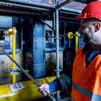 Pracownik w czerwonej kasku i kamizelce ochronnej reguluje przepływ w przemysłowej instalacji w modernizowanej baterii koksowniczej JSW KOKS w Dąbrowie Górniczej. (wygenerowano przez AI)