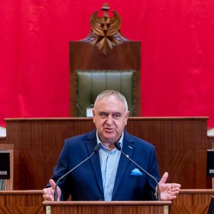 Mężczyzna w ciemnym garniturze przemawia z podium w sali Sejmu Śląskiego, omawiając finansowanie rodzinnych ogrodów działkowych. (wygenerowano przez AI)