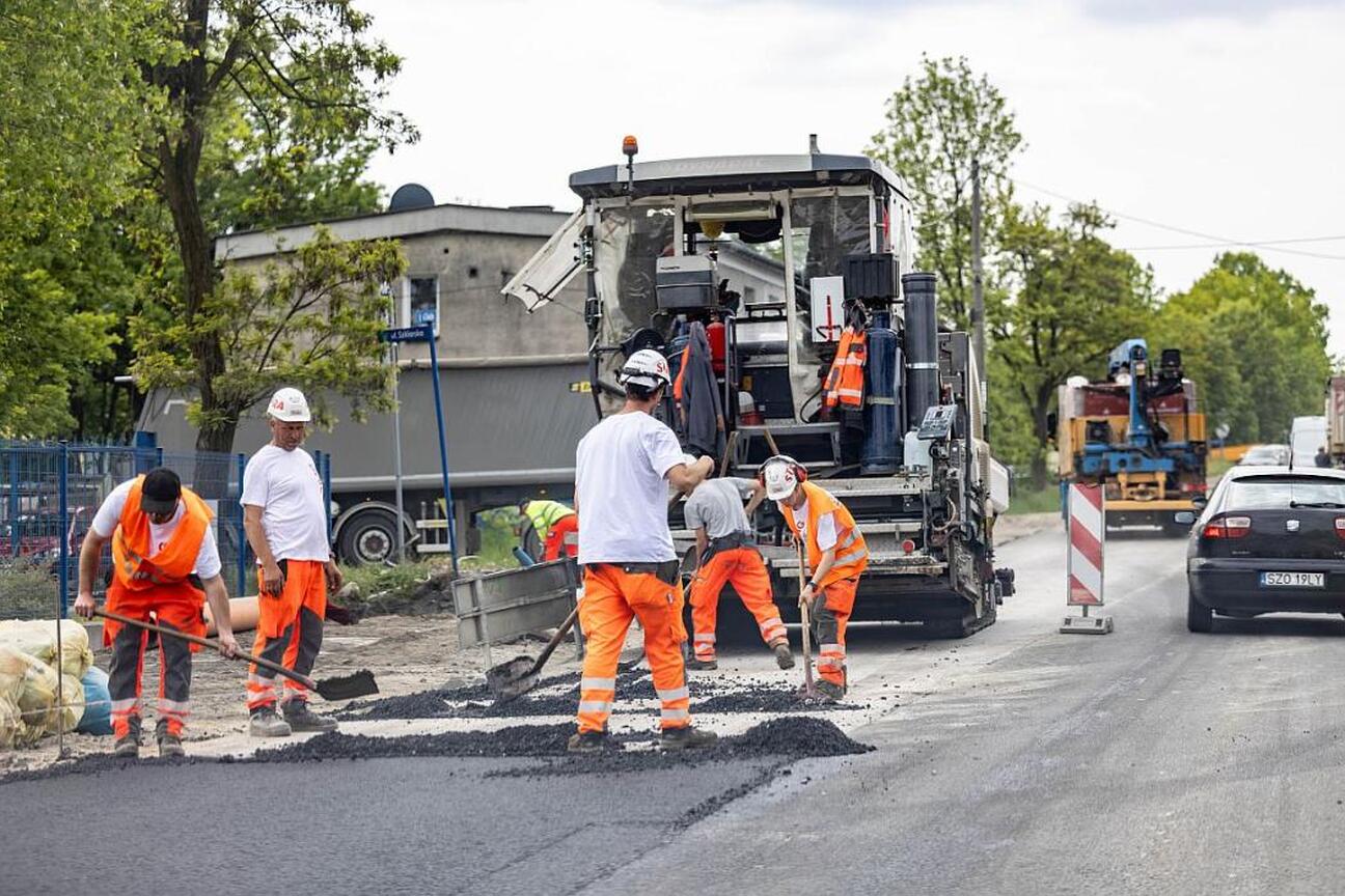 Pracownicy budowlani w kamizelkach ochronnych i hełmach rozprowadzają asfalt na nowo wybudowanej drodze. W tle widoczny jest maszyna do asfaltowania. (wygenerowano przez AI)