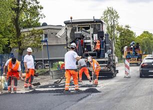 Pracownicy budowlani w kamizelkach ochronnych i hełmach rozprowadzają asfalt na nowo wybudowanej drodze. W tle widoczny jest maszyna do asfaltowania. (wygenerowano przez AI)