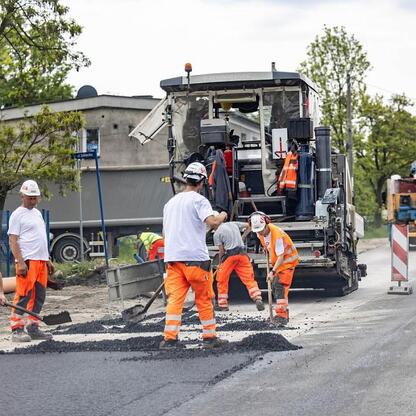 Pracownicy budowlani w kamizelkach ochronnych i hełmach rozprowadzają asfalt na nowo wybudowanej drodze. W tle widoczny jest maszyna do asfaltowania. (wygenerowano przez AI)