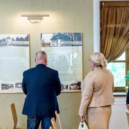 Grupa ludzi w strojach formalnych obserwuje tablice informacyjne w sali z oknami, związana z inicjatywą lokalną. (wygenerowano przez AI)