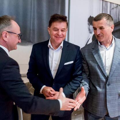 Trzech mężczyzn w strojach formalnych, w trakcie ceremonii, wymieniają uściski rąk. W tle widoczna scena z dużym obrazem i tekstem, sugerując formalny akt. (wygenerowano przez AI)