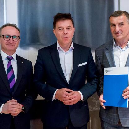 Trzech mężczyzn w garniturach stojących przed plakatem, jednego z nich Grzegorz Boski, wicemarszałek województwa, wręcza dokumenty. (wygenerowano przez AI)