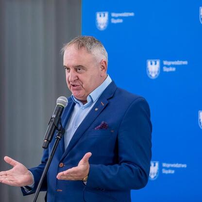 Mężczyzna w niebieskim garniturze przemawia z podium w Województwie Śląskim, podczas uroczystości przekazania umów na modernizację dróg dojazdowych do terenów wiejskich. (wygenerowano przez AI)