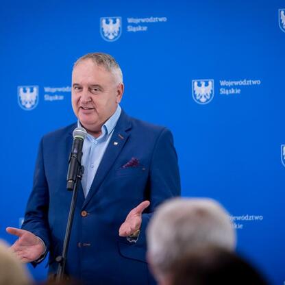 Mężczyzna w niebieskim garniturze przemawia na podium w Województwie Śląskim, podczas uroczystości przekazania umów na modernizację dróg dojazdowych do terenów wiejskich. (wygenerowano przez AI)