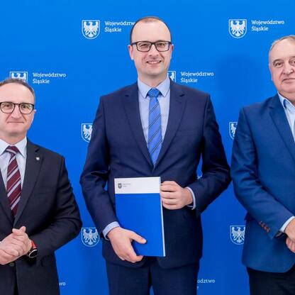 Trzech mężczyzn w strojach formalnych przed emblematem i napisem 