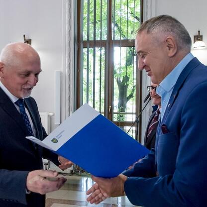 Dwa mężczyźni w strojach formalnych wymieniają niebieski folder w pomieszczeniu z dużymi oknami, oznaczonym jako Biuro Marszałka Województwa Śląskiego. (wygenerowano przez AI)