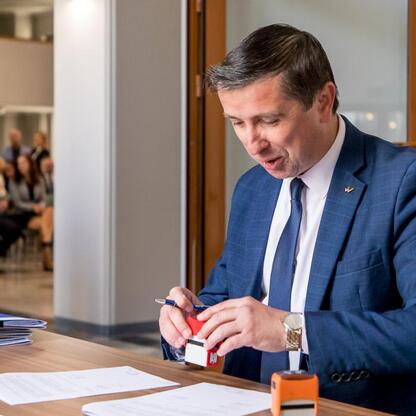 Mężczyzna w niebieskim garniturze podpisuje dokument, stojąc przy biurku z dokumentami i folderami. W tle widoczni ludzie w formalnej sytuacji. (wygenerowano przez AI)
