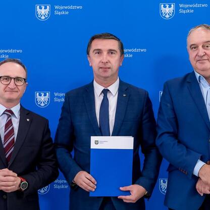 Trzech mężczyzn w garniturach stoi przed emblematem Województwa Śląskiego, jeden trzyma folder. (wygenerowano przez AI)