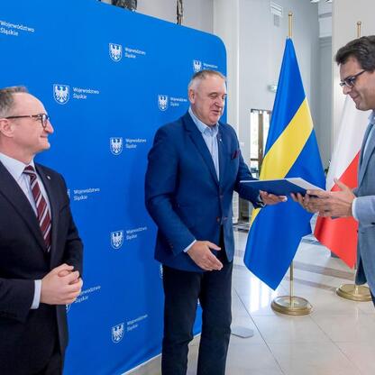 Trzech mężczyzn w strojach urzędniczych stojących przed sztandarem Województwa Śląskiego, podczas ceremonii przekazania umów na modernizację dróg wiejskich. (wygenerowano przez AI)
