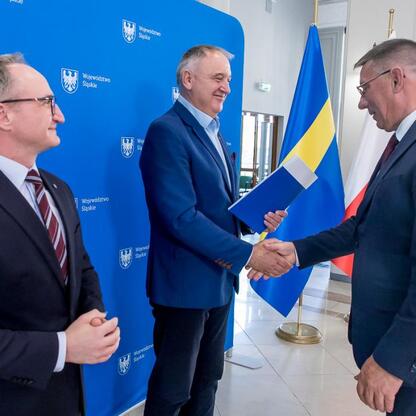 Trzech urzędników w strojach formalnych trzyma się za ręce przed emblematem i nazwą Województwa Śląskiego. (wygenerowano przez AI)