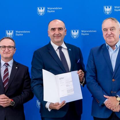 Trzech mężczyzn w strojach urzędniczych przed emblematem Województwa Śląskiego. W centrum stoi Grzegorz Boski, wicemarszałek, trzymający dokument, obok stoi Marek Bieniek, przewodniczący Komisji Rolni (wygenerowano przez AI)