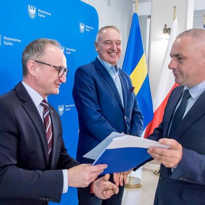 Trzech urzędników w strojach formalnych, którzy podczas uroczystości wręczają umowy na modernizację dróg dojazdowych do terenów wiejskich województwa śląskiego. (wygenerowano przez AI)