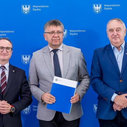 Trzech mężczyzn w strojach formalnych stoi przed niebieskim tłem z emblematem Województwa Śląskiego. W centrum stoi Grzegorz Boski, wicemarszałek, trzymający folder, podczas ceremonii przekazania umów (wygenerowano przez AI)