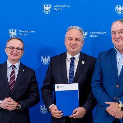 Trzech mężczyzn w strojach urzędniczych przed emblematem Województwa Śląskiego. W centrum Grzegorz Boski z folderem, po lewej Marcin Ryś z rękami skrzyżowanymi, po prawej Joanna Pasierbek-Konieczny z  (wygenerowano przez AI)