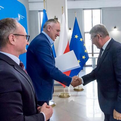 Trzech mężczyzn w strojach urzędniczych trzyma się za ręce w formacji, przedstawiając umowy modernizacji dróg dojazdowych do terenów wiejskich województwa śląskiego. (wygenerowano przez AI)