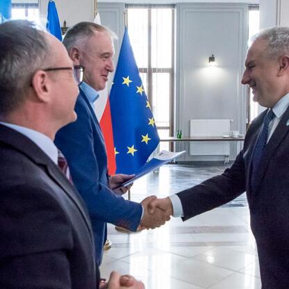 Trzech mężczyzn w strojach formalnych trzyma się za ręce w biurze marszałkowskim województwa śląskiego, podczas ceremonii przekazania umów na modernizację dróg dojazdowych do terenów wiejskich. (wygenerowano przez AI)